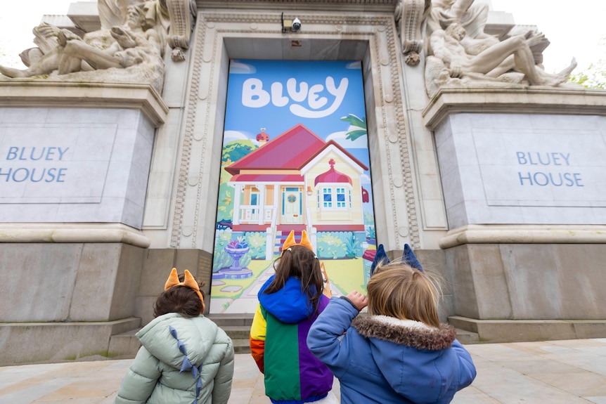 Three children standing in front of Australia House in London, draped with a Bluey banner and signs saying 'Bluey House'.