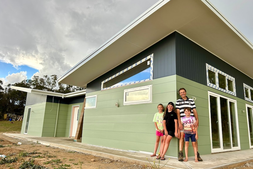 A father and his two daughters and son stand in front of a home on an overcast day.