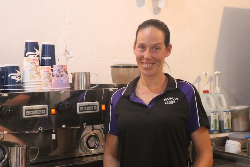 A woman in a black and purple polo shirt smiles as she stands in front of a large coffee machine.