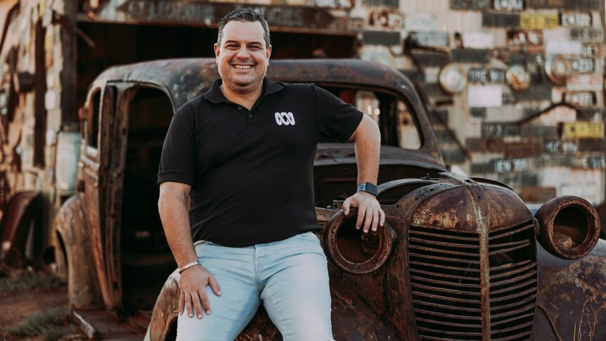 Ivo Da Silva leaning on the hood of a wrecked and rusty classic car, in the middle of a junkyard.