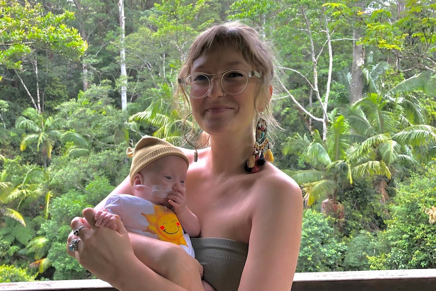 Alana Wilkinson smiling while holding baby Rafferty, trees behind them