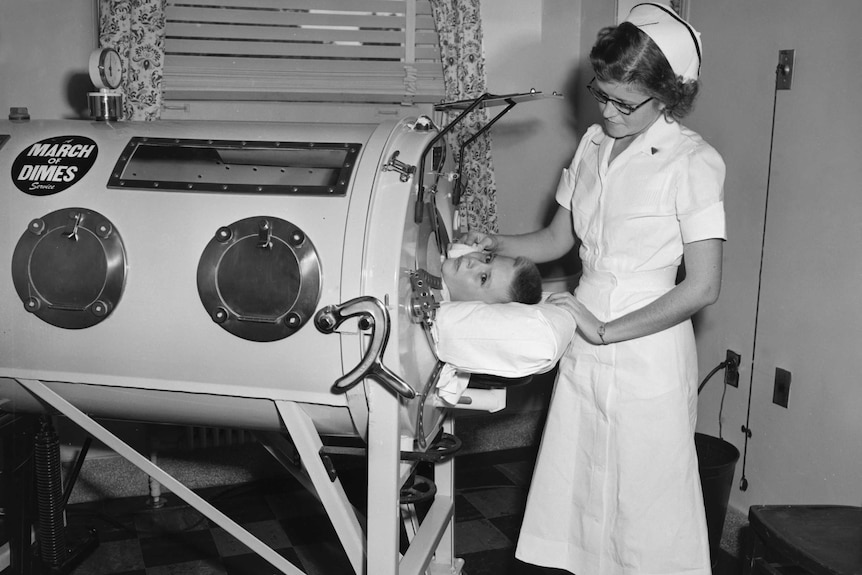 A black and white photo of a young boy lying in an iron lung with a nurse standing near his head.