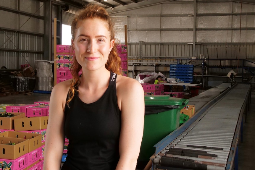 A young woman with her hair up sits next to a conveyor belt in a packing shed.