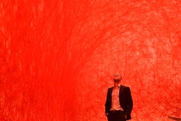 A man in a dark suit stands in an orange room staring up at the ceiling.