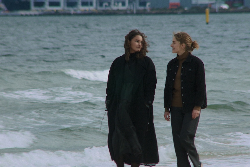 Two women in their mid 20s in dark clothes walking on a beach