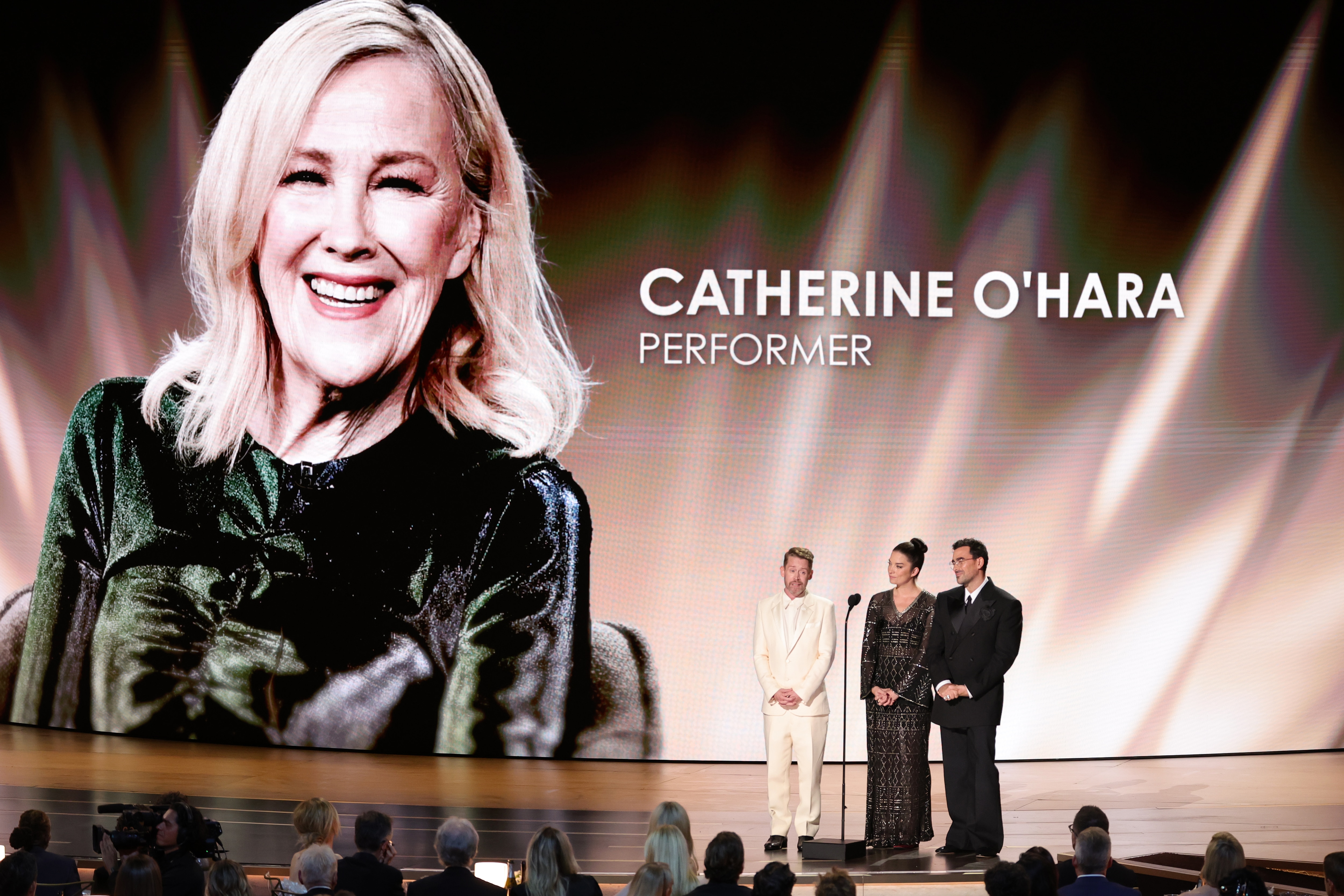 Three people stand on stage with a photo of Catherine O'Hara as tribute on the big screen behind.