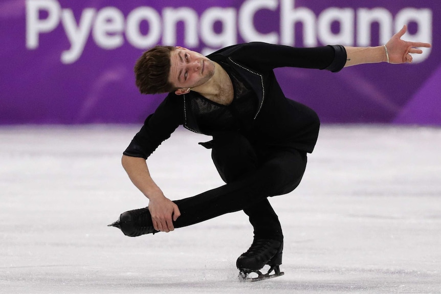 A male figure skater in a black outfit squats down and holds his foot with his hand as he spins