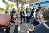 A wide shot showing a media scrum around Anthony Albanese and Mark McGowan sitting down outdoors, with Albanese holding a baby.