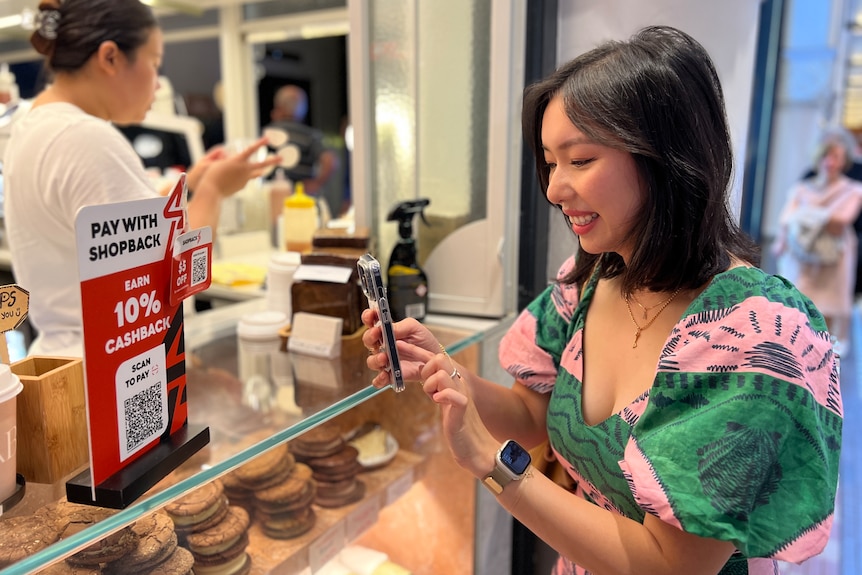 Viona Agung is standing in front of a cafe, holding her smartphone up to capture a QR code displayed on the front counter.