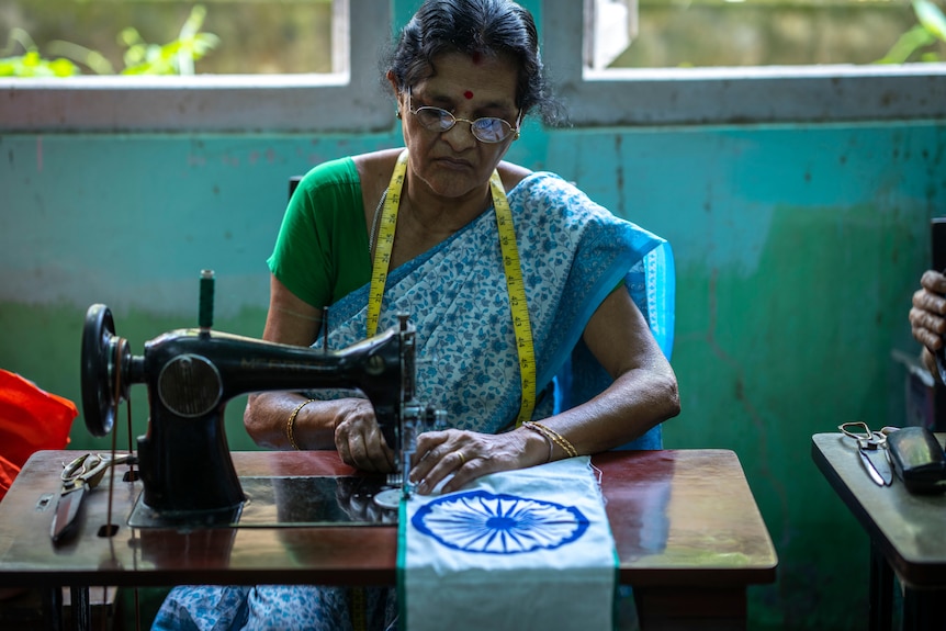 A woman in a blue and green outfit with a bindi sits at a sewing machine with part of the Indian flag. 