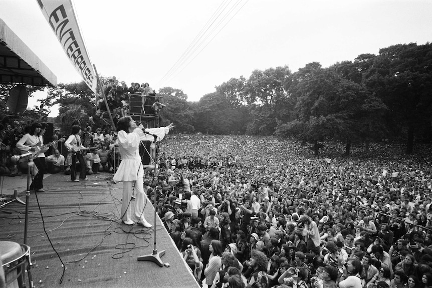 Mick Jagger sings to an enormous crowd with bandmates on stage. He is wearing a vintage smock top
