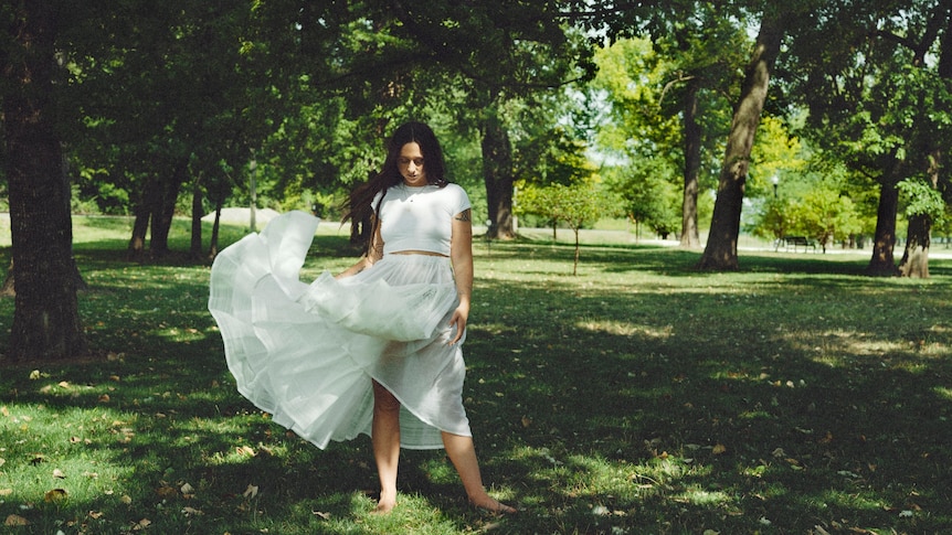 A woman in a white dress standing in a leafy park. The wind blows the dress up.