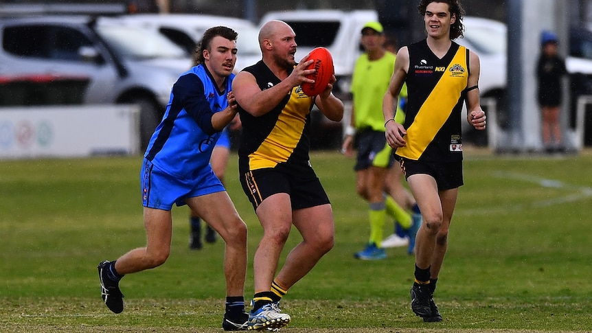 A man wearing a yellow and black football jumper holding a ball while a man in a blue jumper stands closely behind, 