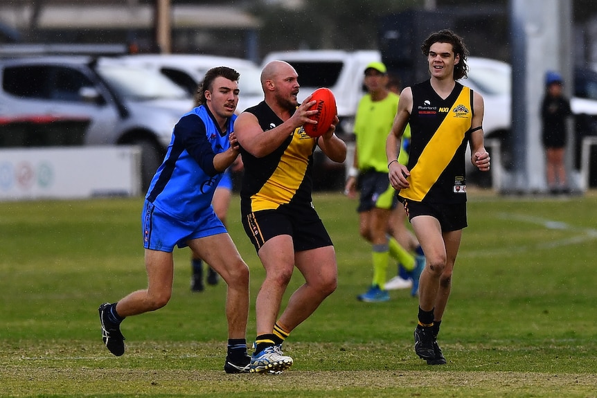 A man wearing a yellow and black football jumper holding a ball while a man in a blue jumper stands closely behind, 