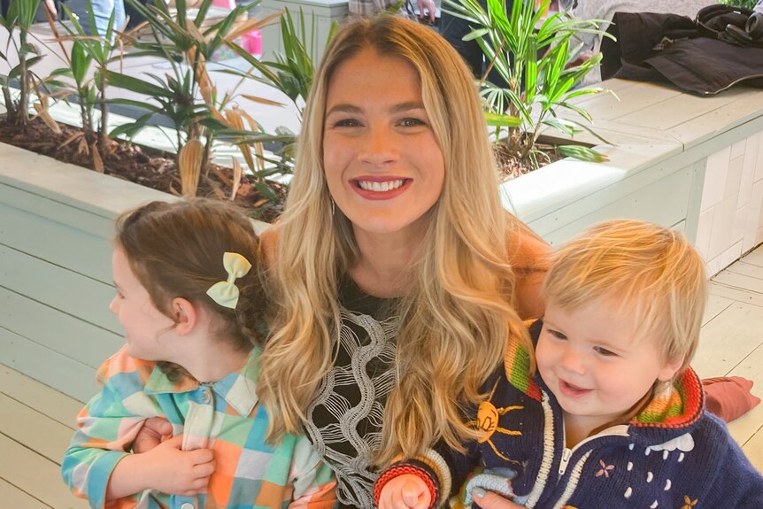 Grace with blonde hair down, smiling at camera with her two young kids on her lap