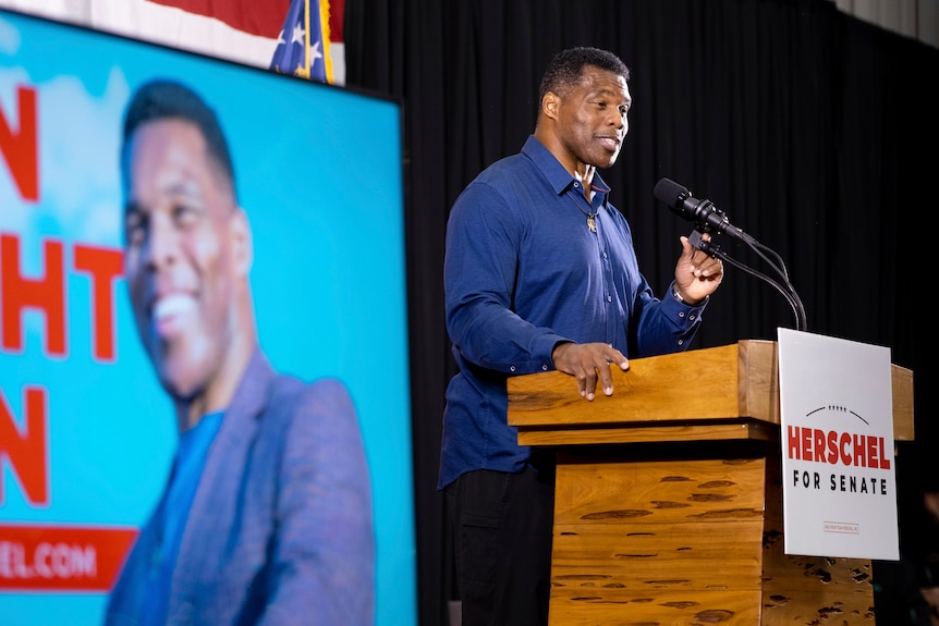 Mr Walker stands speaking at a podium with a large screen showing his face behind him. 