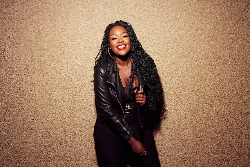 Ruva Ngwenya, a 30-year-old Black woman with long dark curls, wears a black jacket and red lipstick in front of a gold wall