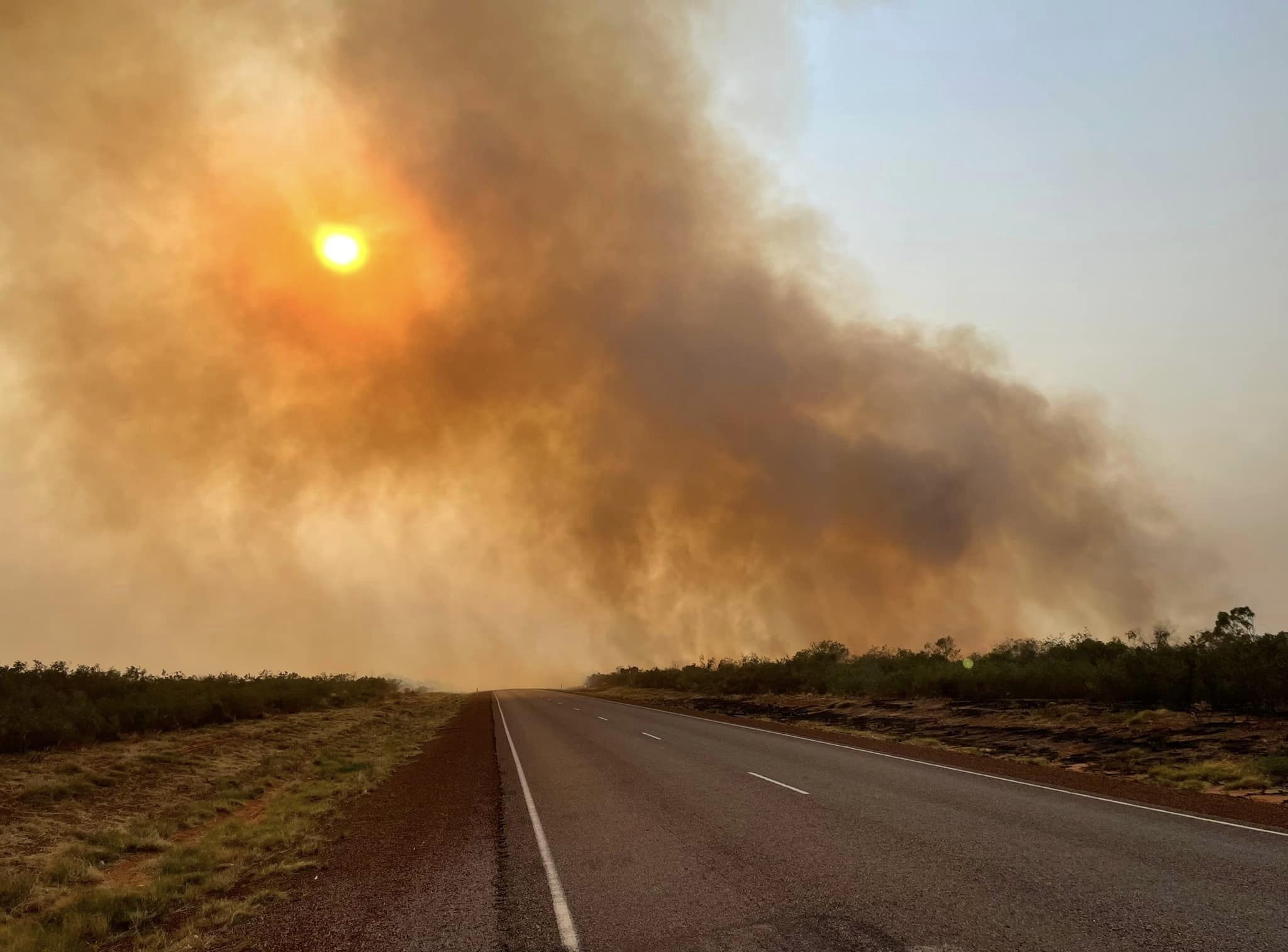 A large cloud of smoke from a bushfire rising into the sky, over a major highway at sunset. 