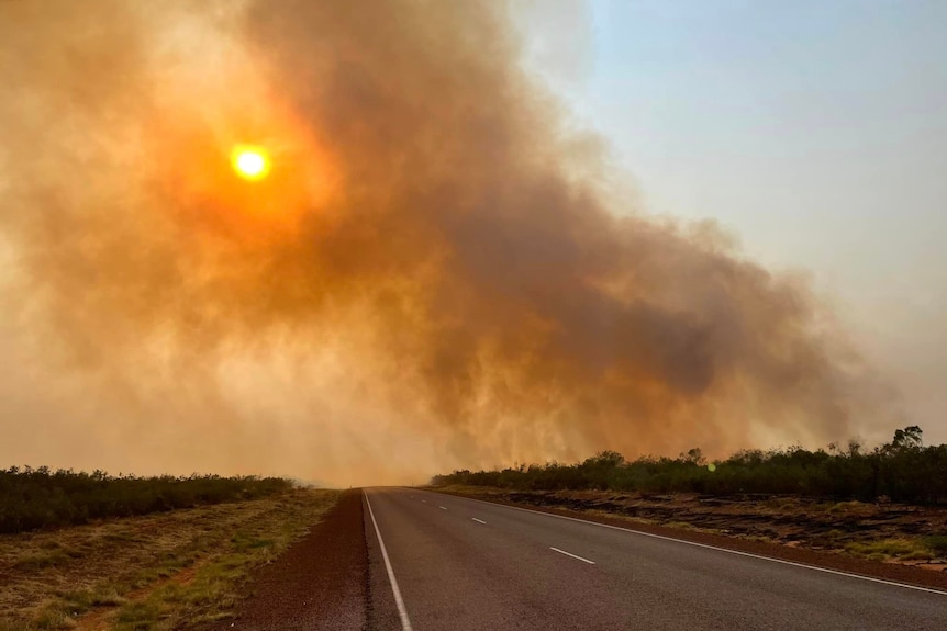 A large cloud of smoke from a bushfire rising into the sky, over a major highway at sunset. 