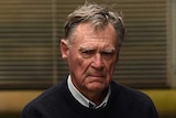 An older man with a stormy expression on his face being led into a police van.