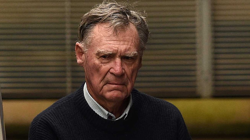 An older man with a stormy expression on his face being led into a police van.