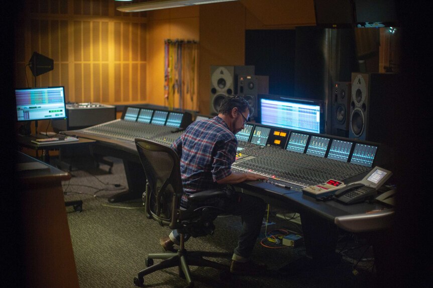 Wales sits in a studio in front of an audio mixing desk.