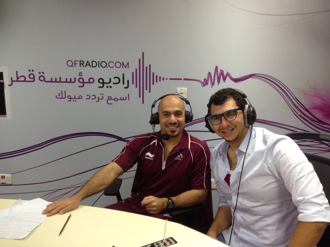 Two men with headsets on sitting at a desk with QFRADIO.COM signage in background.