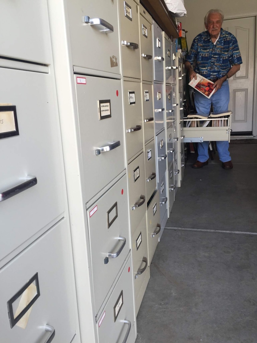 A elderly man holds up a fire safety manual while standing beside a large filing cabinet.