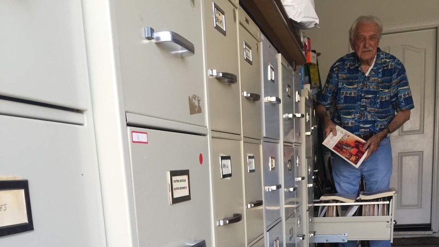 A elderly man holds up a fire safety manual while standing beside a large filing cabinet.