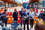 Bill Shorten stands in front of several other politicians and a tram at a press conference.