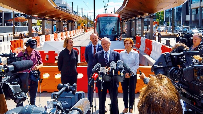 Bill Shorten stands in front of several other politicians and a tram at a press conference.