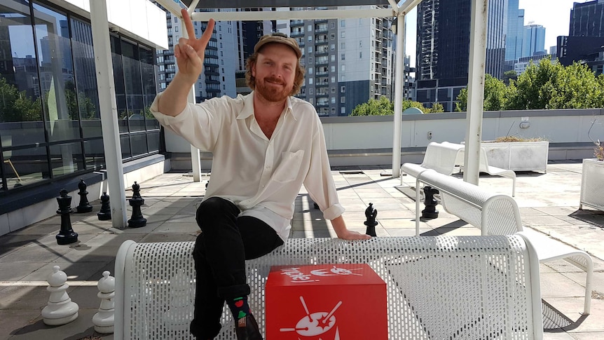 Sam from Jungle Giants sitting on a bench giving a peace sign next to the triple j logo