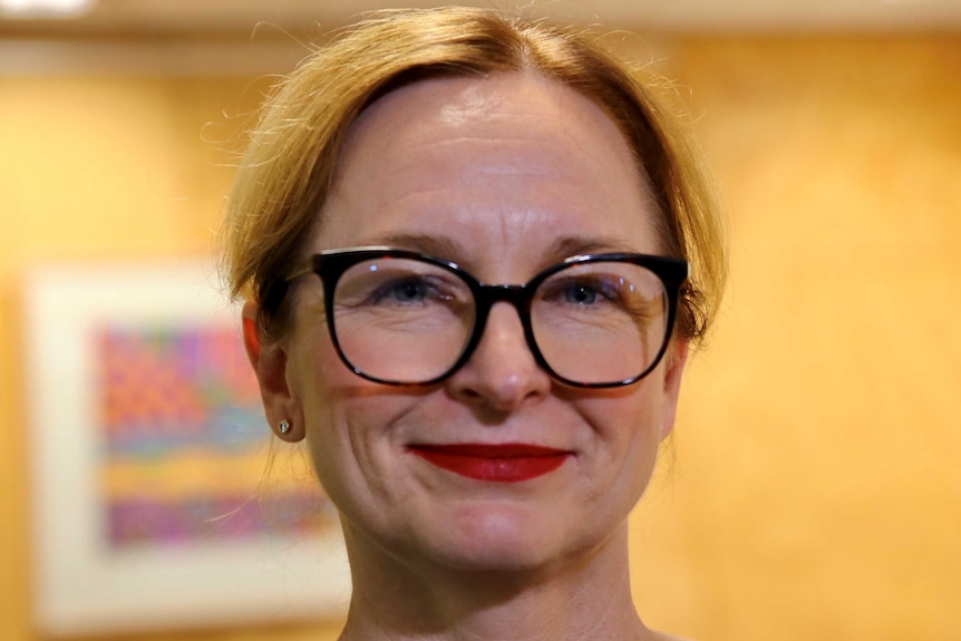 A close up of Lanie, with red lipstick and glasses, with a dark yellow wall behind her.