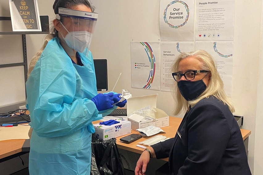 A woman sits in an office, getting prepared to be swabbed by a woman in protective clothing.