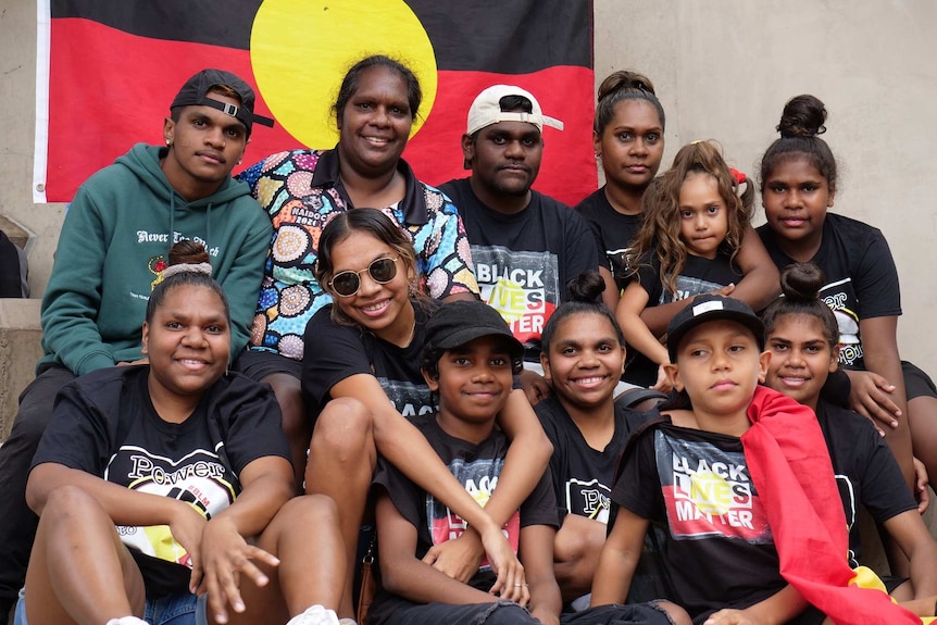 Aunty Kylie Major-Oakley sits amongst a group of young people with an Aboriginal flag in the background.