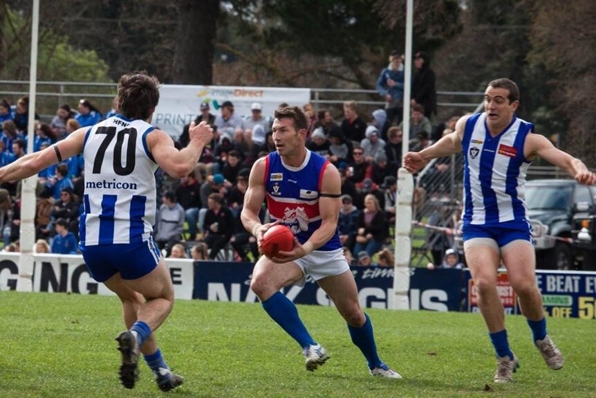 A man in a blue and red football guernsey dodges opponents with ball in hand.