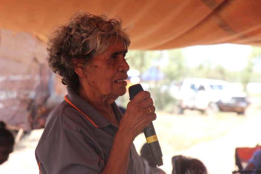 A woman standing under a tent and holding a mic, with other blurred faces sitting on chairs in the background.