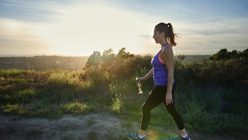 A woman wearing fitness gear and holding a drink bottle walks along a dirt path