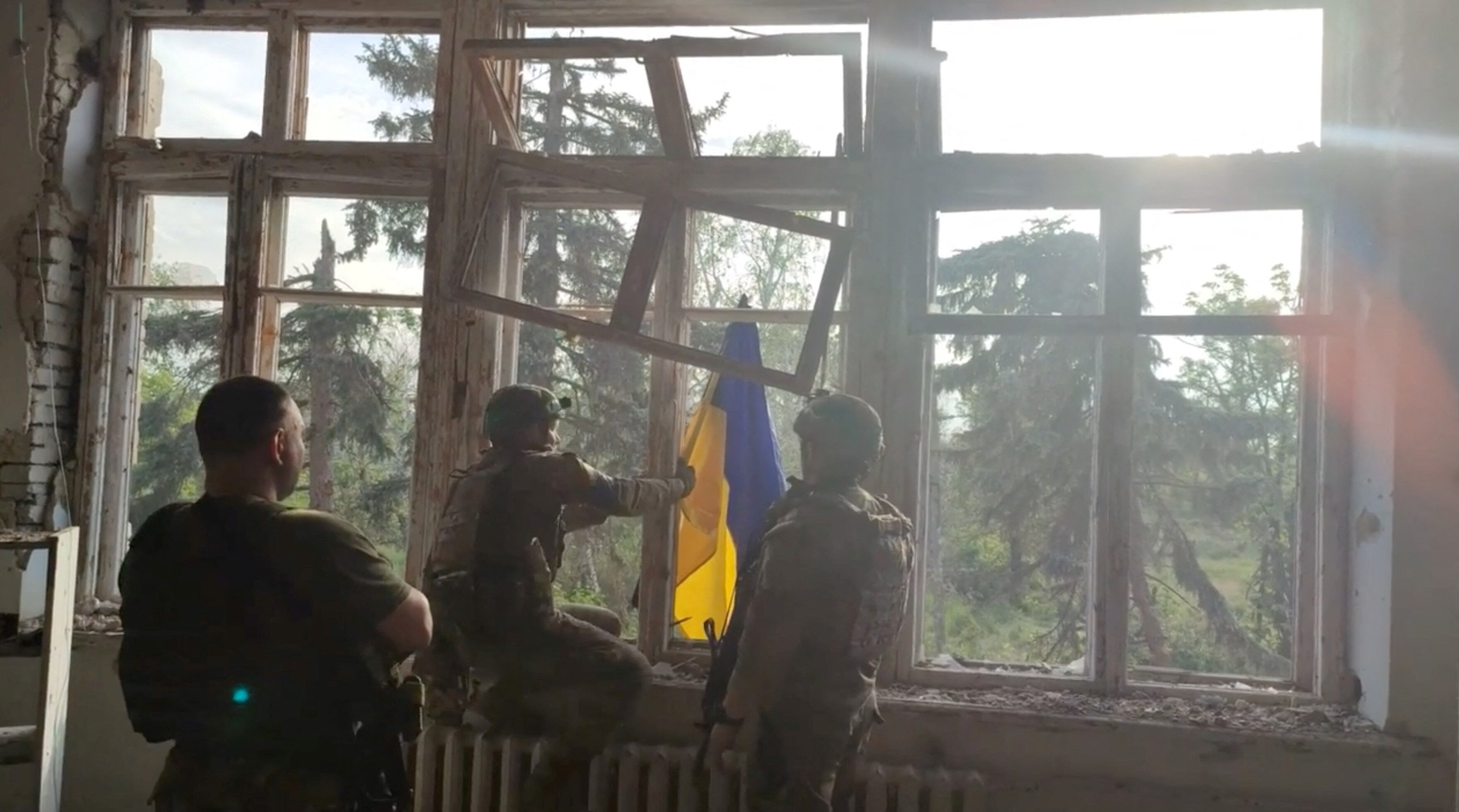 A view of the back of three Ukrainian soldiers placing a Ukraine flag out an open window. 