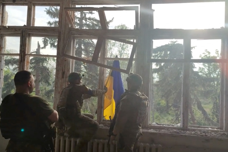 A view of the back of three Ukrainian soldiers placing a Ukraine flag out an open window. 