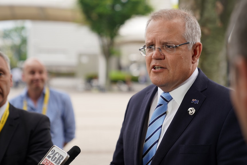 Scott Morrison, wearing a suit with a pale blue tie striped in navy and an Australia flag pin on his lapel, speaks to a reporter