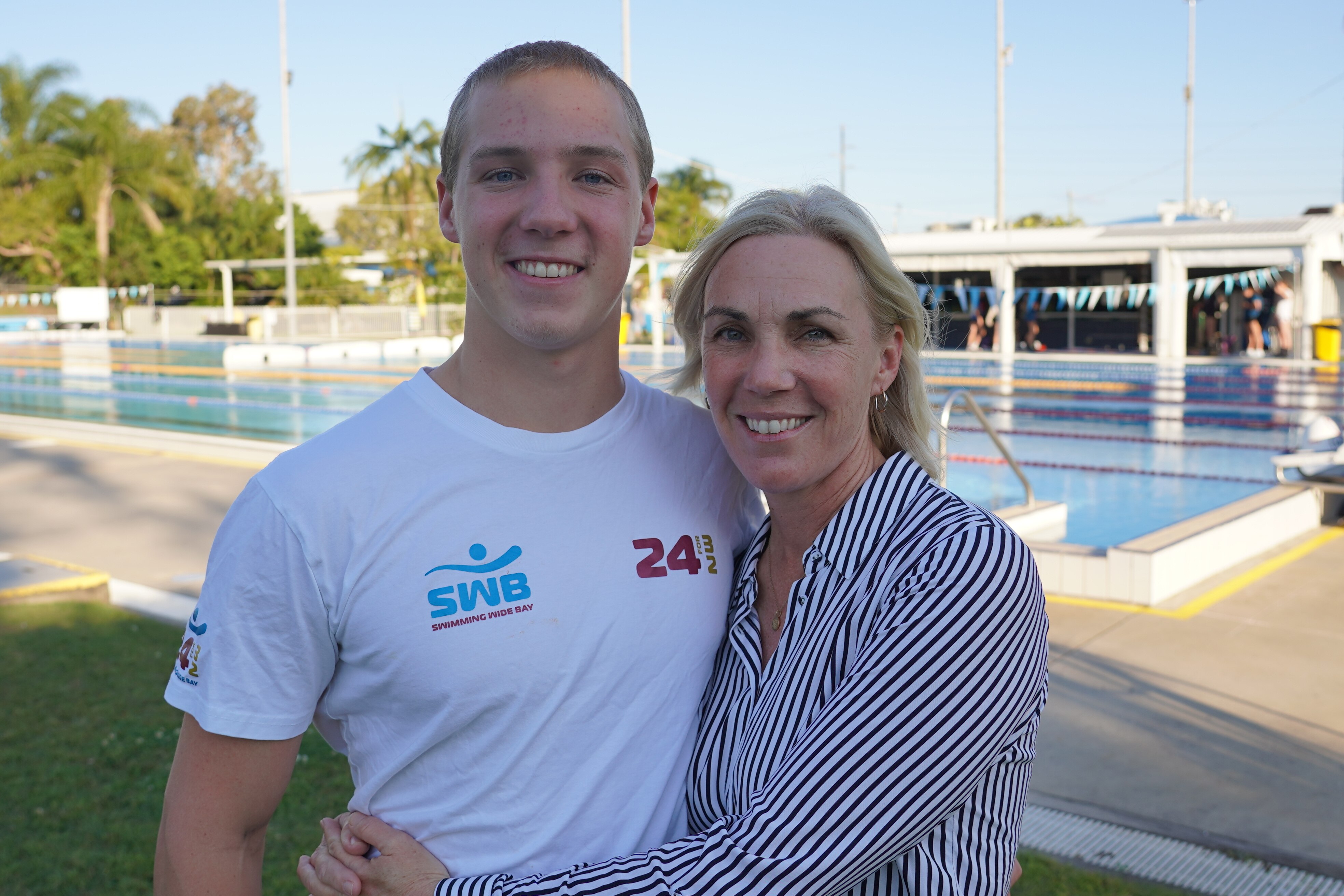 A teenager boy stands next to his mum who has her arms wrapped around his waist in front of a swimming lap pool.