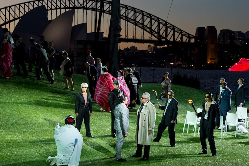 Outdoor stage with green hills and many performers, and silhouette of Sydney Opera House and Harbour Bridge visible in dusk.