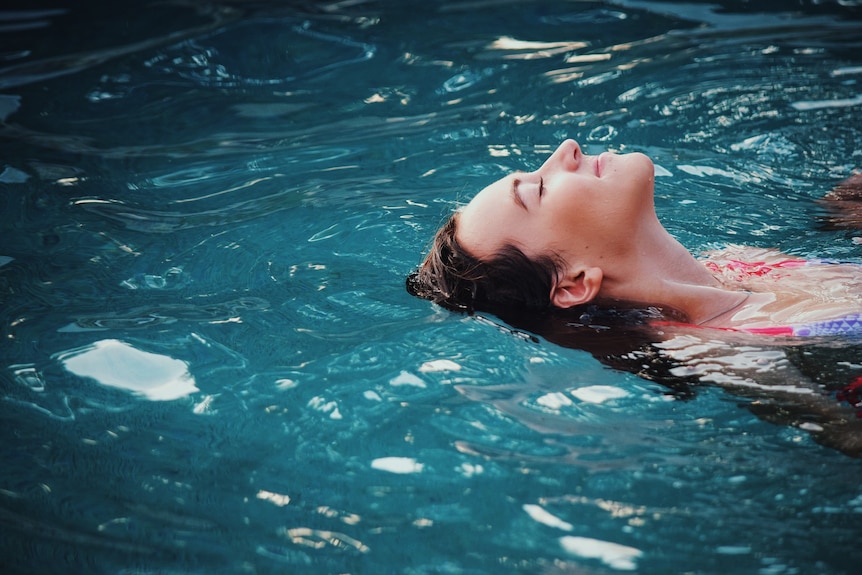A woman floating on her back in a pool with her eyes closed, relaxing