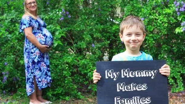 Charli Lee stands in background while heavily pregnant, with her smiling son holding a sign 'my mummy makes families'.