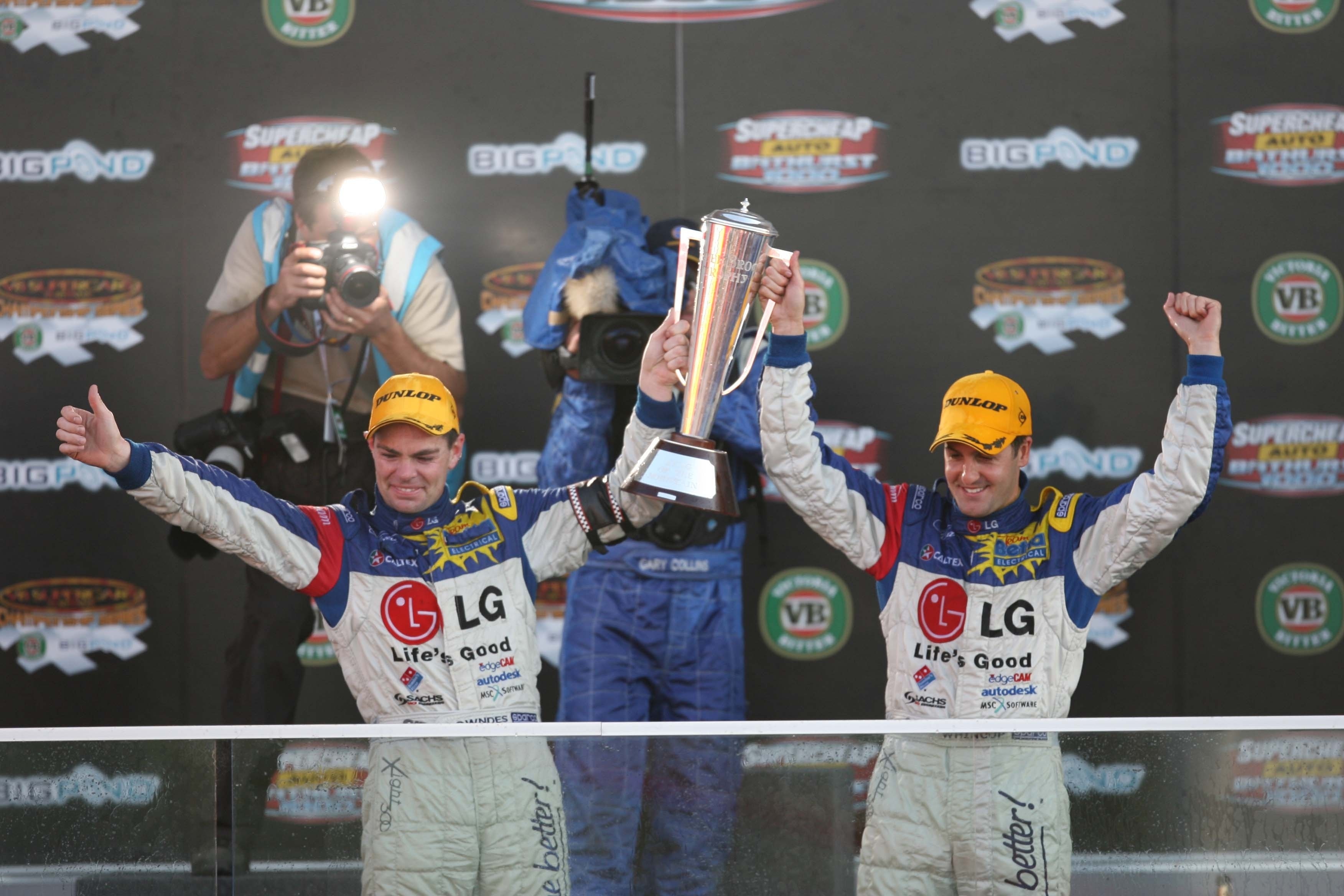 Two drivers standing on the podium, holding the winner's trophy in celebation