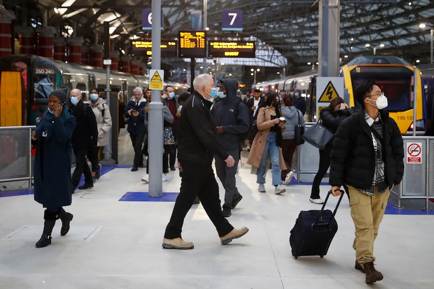 People walk through a busy train station, one man pulling a suitcase behind him, one woman on the phone