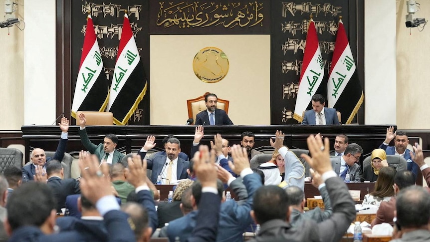 People in suits sitting down with their right hands up voting, with a man sitting at a bench behind them with flags on his side.