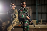 A female Australian soldier and male Indonesian soldier, wearing camouflage, holding guns, leaning against a truck, smiling.