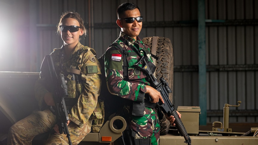 A female Australian soldier and male Indonesian soldier, wearing camouflage, holding guns, leaning against a truck, smiling.